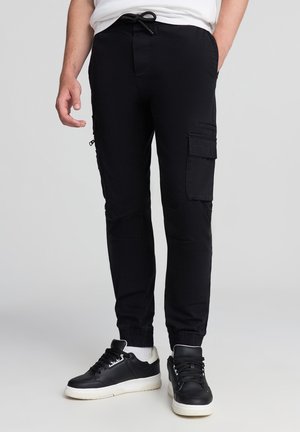 Pantaloni cargo neri in cotone con tasche laterali, polsini elasticizzati e vita con cordino. Indossati con scarpe da ginnastica nere e calzini bianchi.