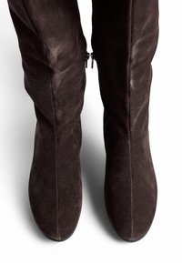 Bottes en daim marron, au-dessus du genou, avec une texture lisse, une forme élancée et des zips sur les côtés pour un accès facile. Design minimaliste, sans quincaillerie visible.
