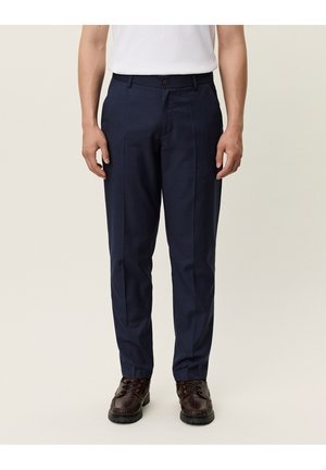 Mann trägt dunkelblaue Stoffhose, dunkelbraune Lederschuhe und ein weißes Hemd und steht vor einem schlichten Hintergrund.