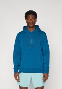 Blå bomulds-hoodie har en frontlomme, ribbede håndled og et cirkulært "Nike Court" logo i kontrastbroderi.