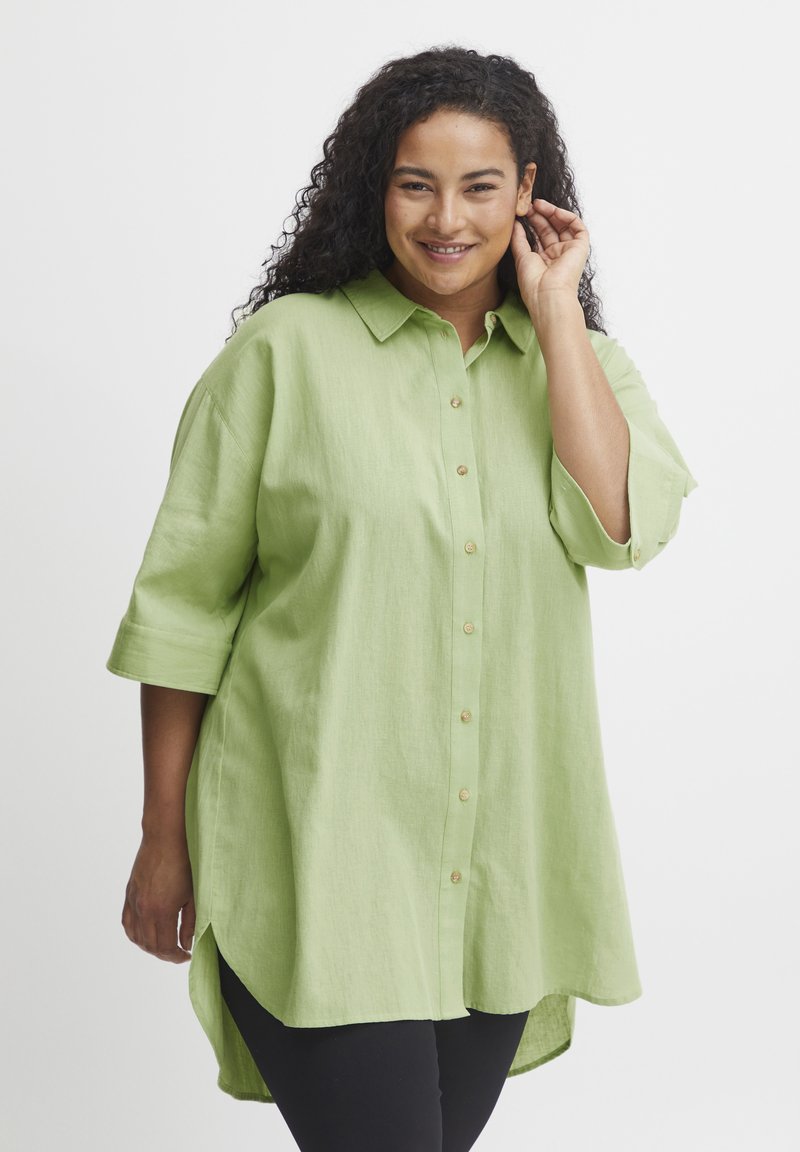 Camicia verde chiaro con bottoni realizzata in un tessuto morbido, con collo a camicia, maniche a tre quarti e un orlo posteriore più lungo.