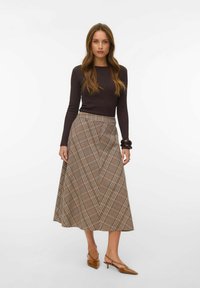 Femme debout devant un fond blanc, portant un haut noir à manches longues, une jupe midi à carreaux marron, des talons slingback marron et des bracelets marron.