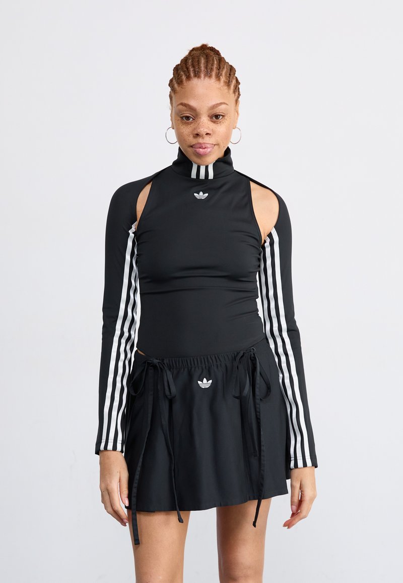 Woman with braided hair wearing a black Adidas cut-out long-sleeve top with white stripes and a matching black skirt with drawstrings.
