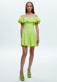 Vestido verde lima con hombros descubiertos, mangas con volantes, botón en la parte delantera y falda con capas. Combinado con sandalias de tacón verdes a juego.