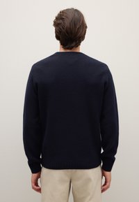 Maglione in maglia testurizzata blu navy con scollo rotondo, polsini e orlo a costine. Indossato sopra pantaloni di colore chiaro, visto da dietro.