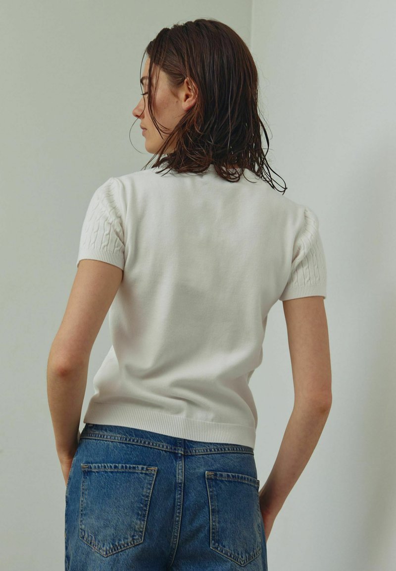 Pull blanc à manches courtes avec épaules en tricot texturé, ourlet côtelé, associé à un jean en denim bleu, vue de dos sur un fond clair.