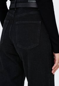 Gros plan sur une personne portant un jean noir taille haute avec des poches arrières et une ceinture noire, la main reposant à côté.