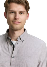 Chemise boutonnée gris clair avec un motif texturé subtil, col sombre, boutons contrastants et un petit logo brodé sur la poitrine.