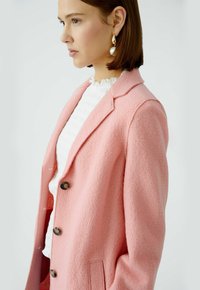 Rosa, strukturierte Blazer mit Revers, braunen Knöpfen und Seiten Taschen, über einer weißen, Rüsche-Bluse geschichtet.