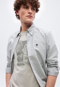 Νεαρός με σγουρά μαλλιά που φοράει ανοιχτό γκρι πουκάμισο με κουμπιά πάνω από μπλουζάκι σε μπεζ χρώμα με γραφικό σχέδιο δακτυλίων δέντρου και τη λέξη "BOSS".