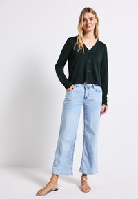 Ciemnozielony kardigan z długim rękawem, z dekoltem w serek i guzikami, zestawiony z jasnoniebieskimi spodniami w stylu wide-leg i srebrnymi sandałami na paski.