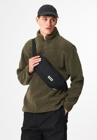 Schwarze Umhängetasche aus strapazierfähigem Stoff mit glatter Textur und Logodetail, quer über der Brust getragen, darüber ein olivgrüner Fleecepullover.