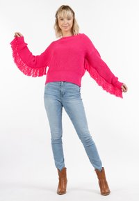 Maglione rosa lavorato a maglia con maniche a frange, abbinato a jeans azzurri e stivaletti marroni. La texture è spessa e vivace.