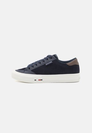 Niski granatowy sneaker z białą podeszwą, brązowym akcentem na pięcie, granatowymi sznurowadłami oraz małą etykietą "Hilfiger" po boku.