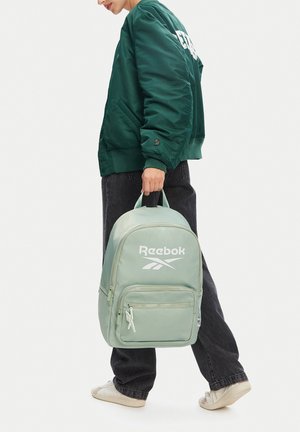 Persona con chaqueta verde y pantalones negros sosteniendo una mochila Reebok verde claro por el asa superior, de pie sobre un fondo blanco.