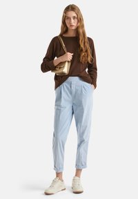 Maglione a maglia marrone, pantaloni a strisce blu chiaro, sneakers color crema. Modello che tiene una borsa dorata. Look casual, vestibilità rilassata con pantaloni risvoltati.