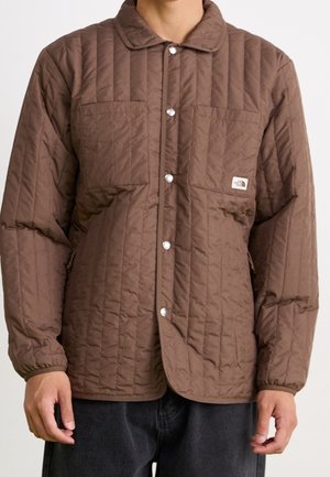 Persoon draagt een bruine gewatteerde jas met drukknopen, twee borstzakken en elastische manchetten, gecombineerd met een donkere broek.