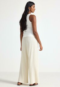 Hvid tanktop parret med løse, teksturerede cremede bukser. Bukserne har et krøllet mønster og cropped længde, suppleret med flade sorte sandaler.