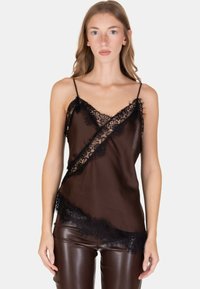 Donna che indossa un camiciaio trasparente con rifiniture in pizzo nero e spalline sottili, abbinato a pantaloni di pelle marrone scuro, in piedi davanti a uno sfondo neutro.