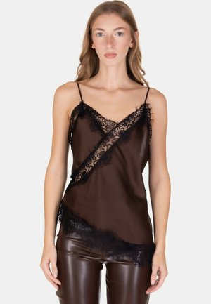 Donna che indossa un camiciaio trasparente con rifiniture in pizzo nero e spalline sottili, abbinato a pantaloni di pelle marrone scuro, in piedi davanti a uno sfondo neutro.