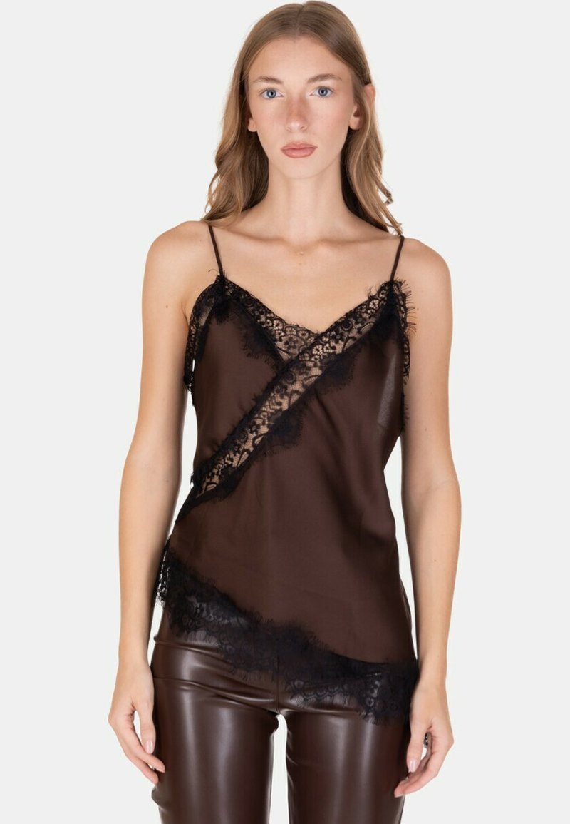 Donna che indossa un camiciaio trasparente con rifiniture in pizzo nero e spalline sottili, abbinato a pantaloni di pelle marrone scuro, in piedi davanti a uno sfondo neutro.