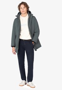 Joven vestido con una chaqueta de invierno con capucha verde, sudadera blanca, pantalones oscuros y zapatillas blancas, de pie con una mano en el bolsillo.