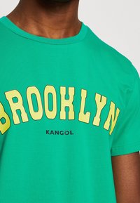 Camiseta verde con gran texto amarillo "BROOKLYN" y un logotipo negro más pequeño de "KANGOL", que lleva puesta una persona de tono de piel medio oscuro.