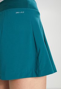 Falda deportiva verde azulado con pliegues, llevada en la cintura y el muslo superior, con el texto blanco "DRI-FIT" en la parte trasera de la cintura.