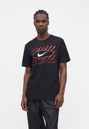 Mladý muž s vrkočmi nosí čierne tričko Nike s červenými diagonálnymi pruhmi a veľkým bielym logom Swoosh, v kombinácii s čiernymi nohavicami.