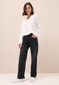 Witte blouse met lange mouwen, een V-hals en knoopdetails, gecombineerd met zwarte rechte jeans en witte sneakers op een neutrale achtergrond.