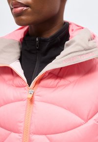 Gros plan d'une personne portant une veste matelassée rose avec un col beige et "KARI TRAA" brodé, sur un haut noir zippé.