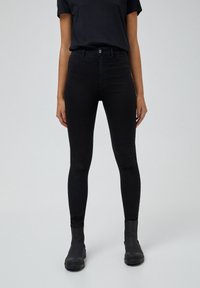 Jeans skinny negros de talle alto, confeccionados en denim elástico, con un diseño clásico de cinco bolsillos y una textura suave, combinados con botas negras.