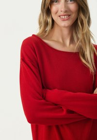 Pull en tricot rouge avec uncol dégage et manches longues, présentant une texture douce et une coupe décontractée. Pas de motifs ni d'accents visibles.