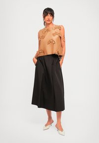 Ženska s tatuji nosi bež brezrokavnik z loki, črne široke culotte hlače in bele mule s koničastim petam, stoji ob enobarvni ozadju.