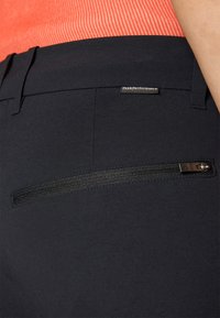 Black pants featuring a zippered pocket on the back and a fabric tag with "PeakPerformance." Smooth texture and tailored fit.