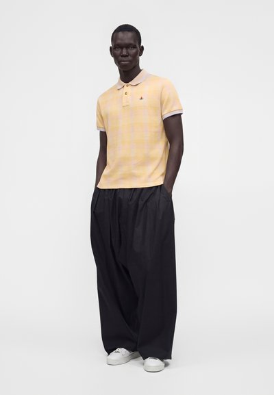 Homme debout, les mains dans les poches, portant un polo jaune à manches courtes, un pantalon noir large et des baskets blanches sur un fond uni.