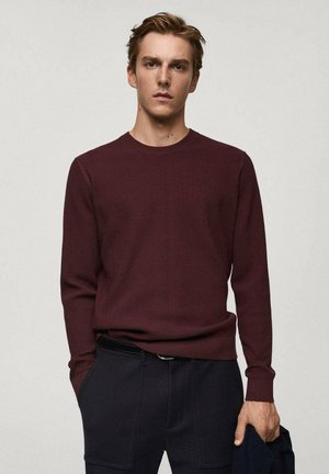 Burgunderfarbener, gerippter Pullover mit langen Ärmeln und Rundhalsausschnitt, aus strukturiertem Stoff gefertigt. Das Modell trägt marineblaue Hosen und hat die Hände in den Taschen.