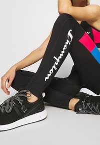 Černé legíny s bílým logem, které mají barevné bloky v růžové a modré. Doplněné černými sportovními botami s bílými podrážkami a vzorovanými tkaničkami.