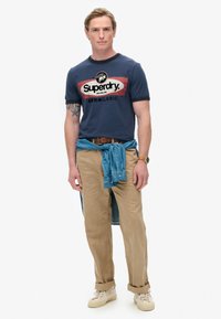 Camiseta navy con estampado de logo en rojo y blanco, combinada con pantalones beige y una camisa de mezclilla atada. Modelo lleva zapatillas beige y un reloj.