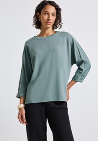 Kvinna med kort lockigt hår som bär en lös teal långärmad tröja med vita ränder på ärmarna och svarta byxor, står mot en enhetlig bakgrund.