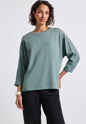 Vrouw met kort krullend haar, gekleed in een losse teal lange mouwen top met witte strepen op de mouwen en zwarte broek, staande tegen een effen achtergrond.