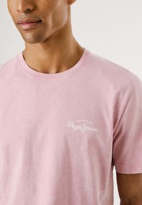 Camiseta de algodón rosa con cuello redondo y mangas cortas, que presenta un logo bordado en blanco en el pecho, con un acabado texturizado.