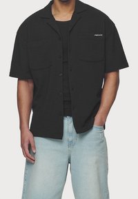 Camicetta nera a maniche corte con finitura strutturata, due tasche sul petto e chiusura con bottoni sul davanti. Indossata sopra una canottiera nera e abbinata a jeans azzurri.