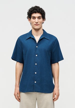 Άνδρας με σγουρά σκούρα μαλλιά, φορώντας μπλε πουκάμισο με κοντά μανίκια και ανοιχτό μπεζ παντελόνια, στέκεται μπροστά σε ένα απλό λευκό φόντο.