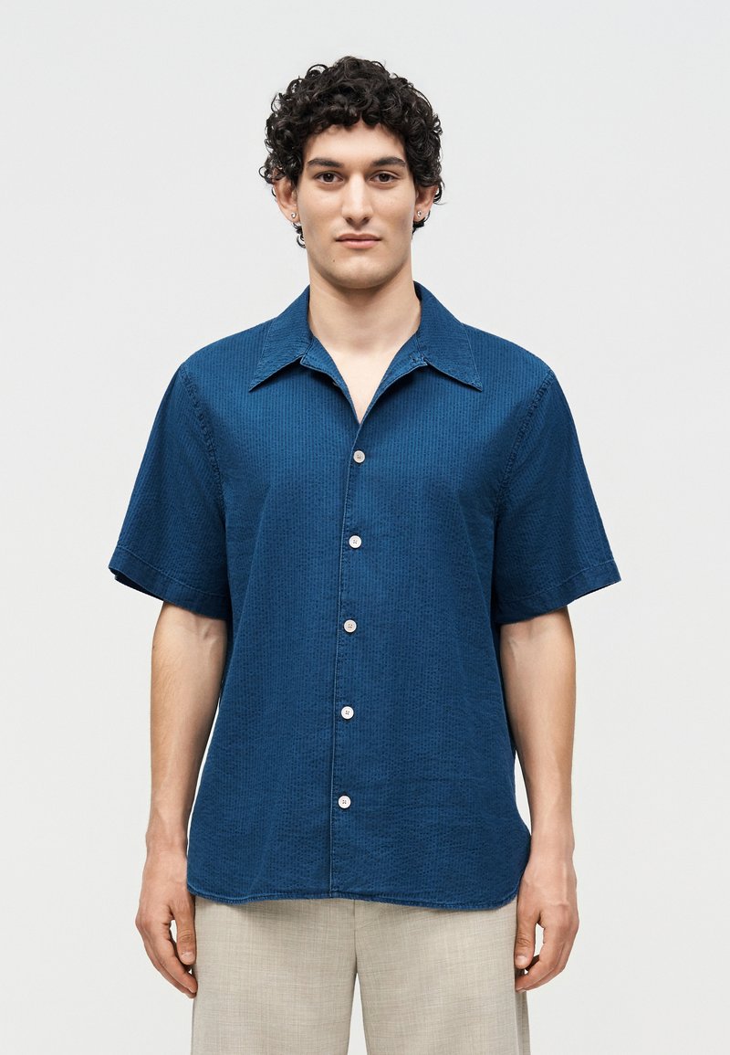 Άνδρας με σγουρά σκούρα μαλλιά, φορώντας μπλε πουκάμισο με κοντά μανίκια και ανοιχτό μπεζ παντελόνια, στέκεται μπροστά σε ένα απλό λευκό φόντο.