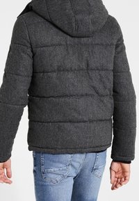 Chaqueta acolchada de tela gris oscuro con capucha. Presenta secciones acolchadas y un corte recto, acompañada de jeans de mezclilla azules.