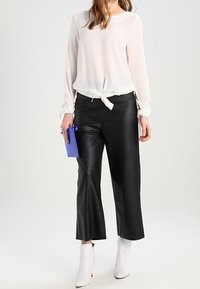 Blusa blanca de manga larga con lazo en la cintura, combinada con pantalones anchos negros de cuero sintético y botines blancos. Sosteniendo un bolso de mano azul.