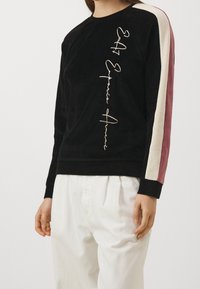 Persona che indossa una felpa nera con scritte beige verticali e strisce rosa e beige sulle maniche, abbinata a pantaloni bianchi larghi.