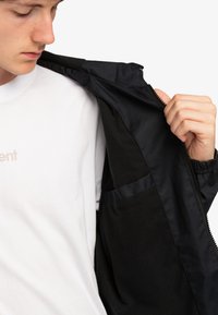 Joven en camiseta blanca mostrando el interior de una chaqueta negra desabrochada con un bolsillo interior, contra un fondo blanco.