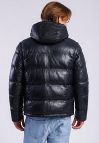 Man met bruin haar die een zwart gewatteerd lederen jack met capuchon draagt, van achteren zichtbaar, gecombineerd met lichtblauwe spijkerbroek.